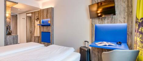 A hotel room with a bed, a desk with a chair, a TV mounted on the wall, and a suitcase on the floor.