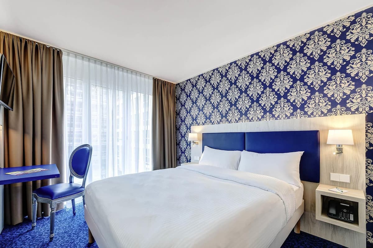 A hotel room with a large bed, a desk, a chair, a TV, and a nightstand with a lamp.