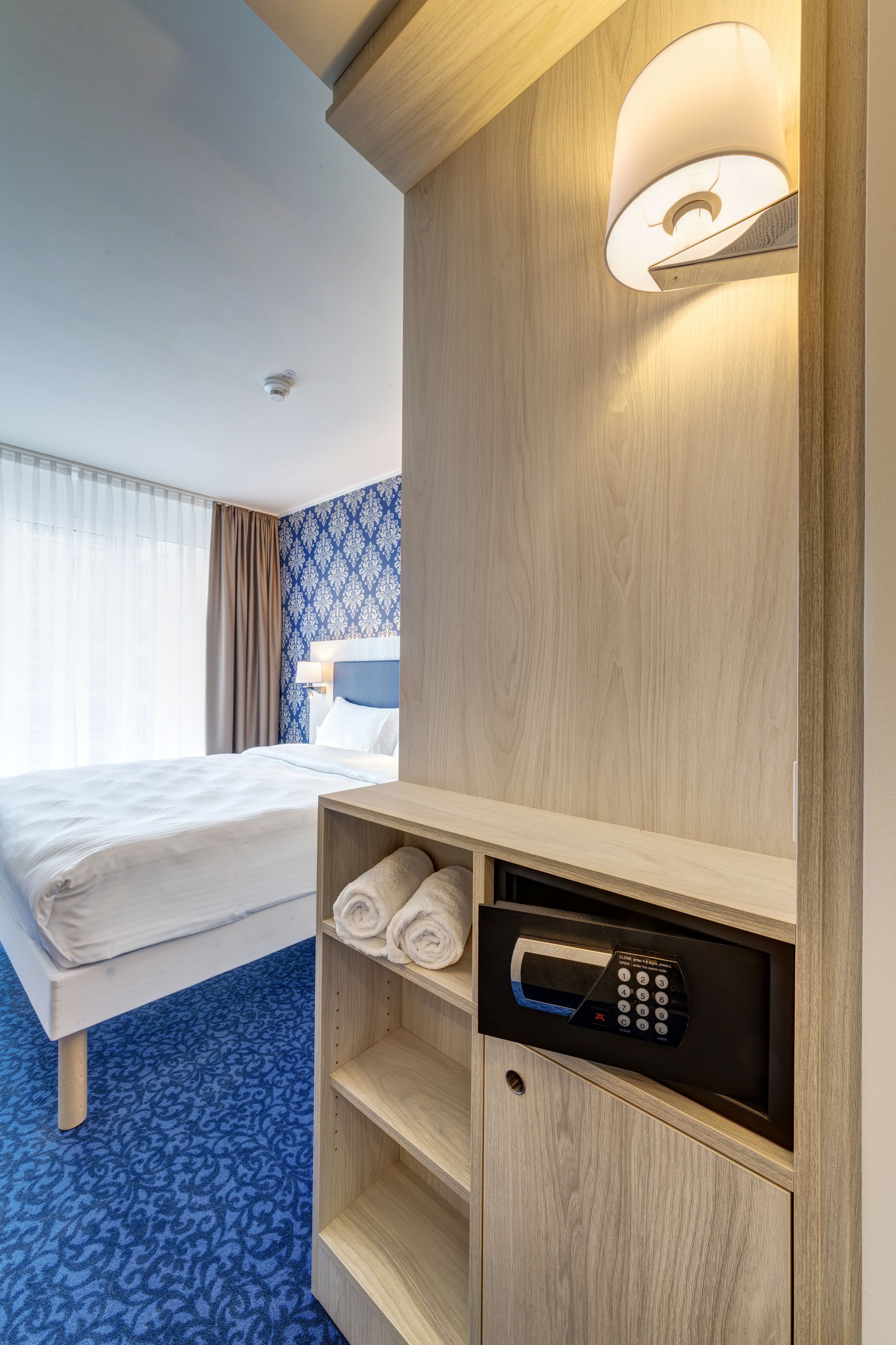 Habitación de hotel con cama, lámpara de pared, microondas integrado y alfombra azul estampada.