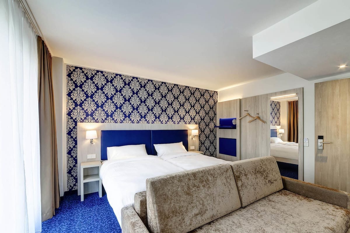 A hotel room with a large bed, a sofa, a nightstand, and a mirror.