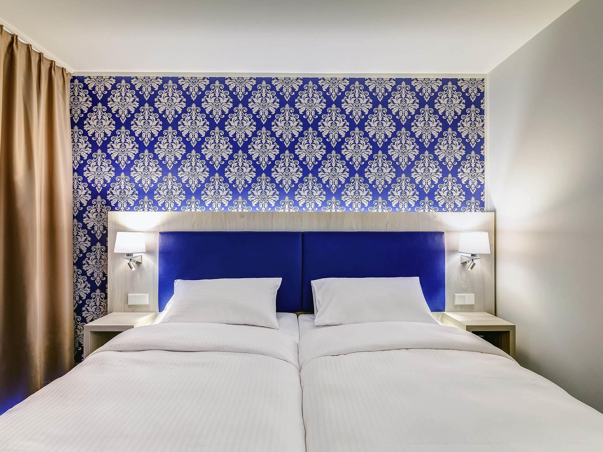 A double bed with a blue headboard, two bedside lamps, and a patterned wallpaper.