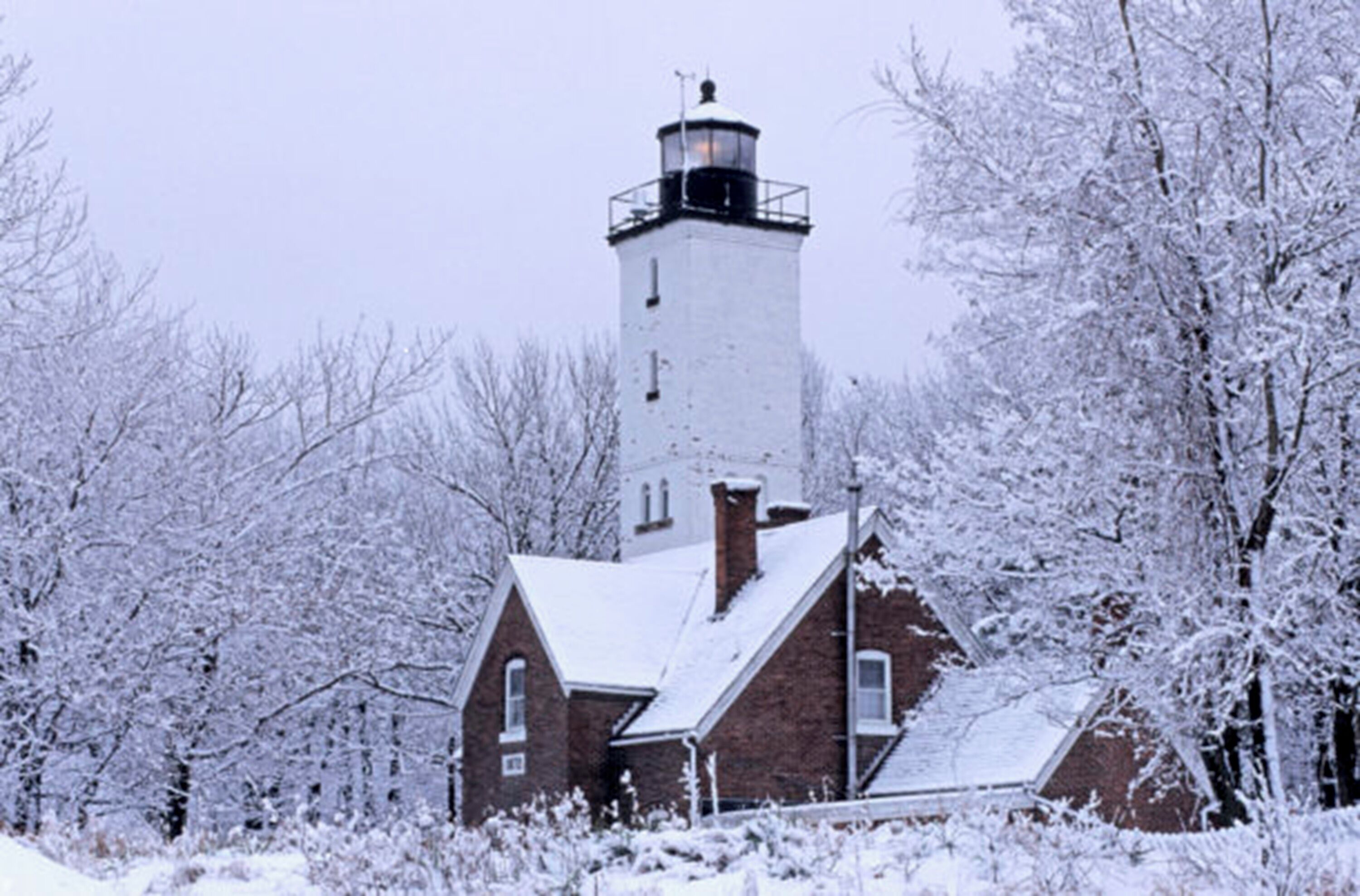 Un faro con un edificio de ladrillo en primer plano, rodeado de árboles cubiertos de nieve.
