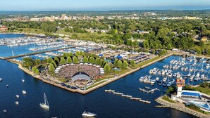 Vista aérea de un puerto deportivo con varias muellas y barcos, rodeado de zonas verdes y edificios urbanos.