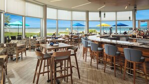 Zona de bar moderna con una barra curva, mobiliario de madera y grandes ventanales con vistas a la playa.