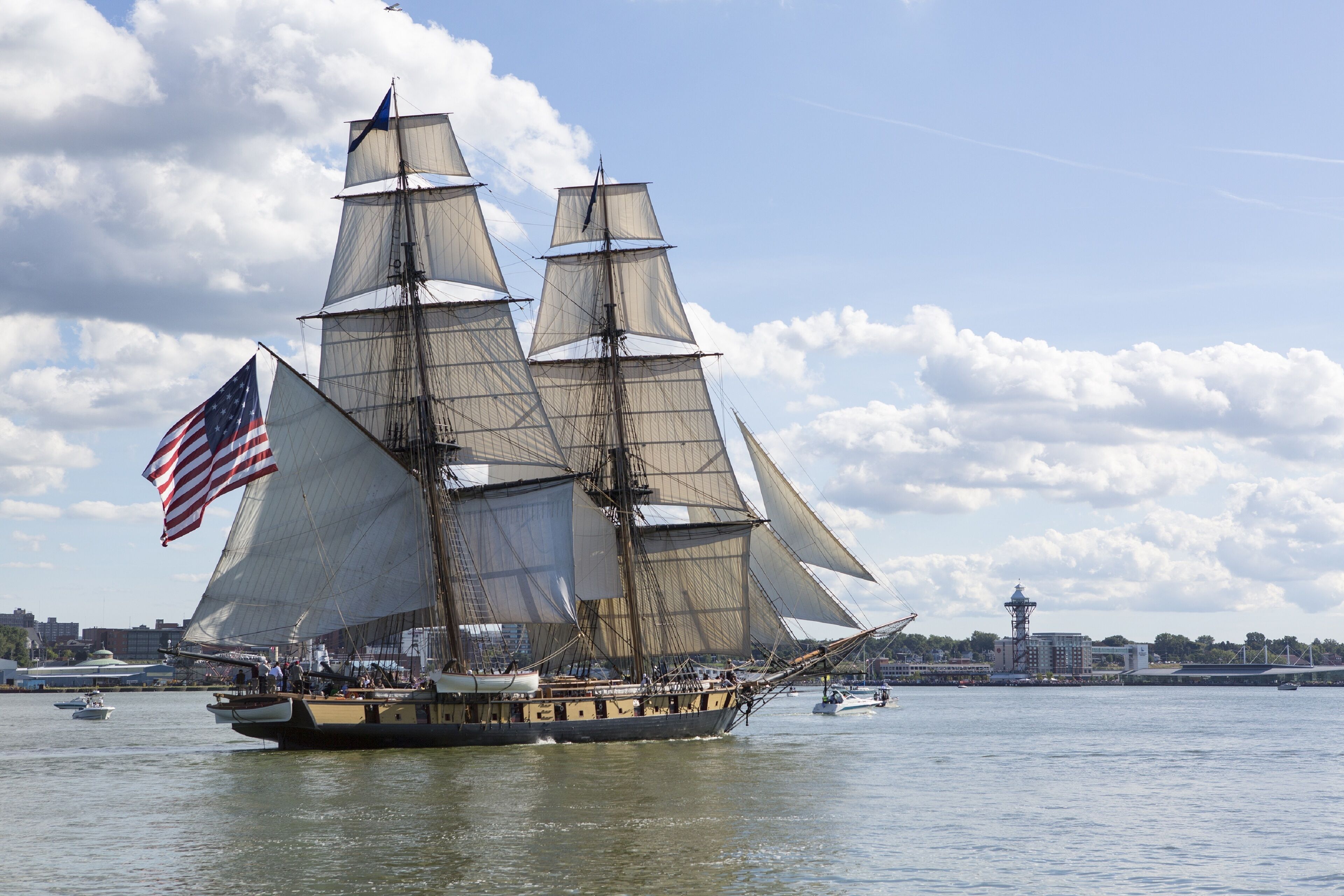 A large sailing ship with multiple masts and sails, docked at a harbor with a cityscape in the background.