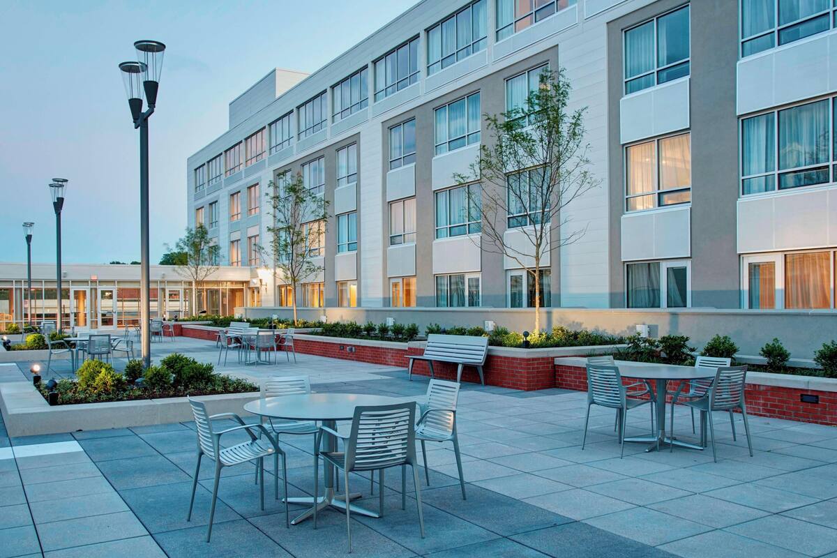 A modern building with multiple floors, outdoor seating area with tables and chairs, and a landscaped garden with small trees and shrubs.