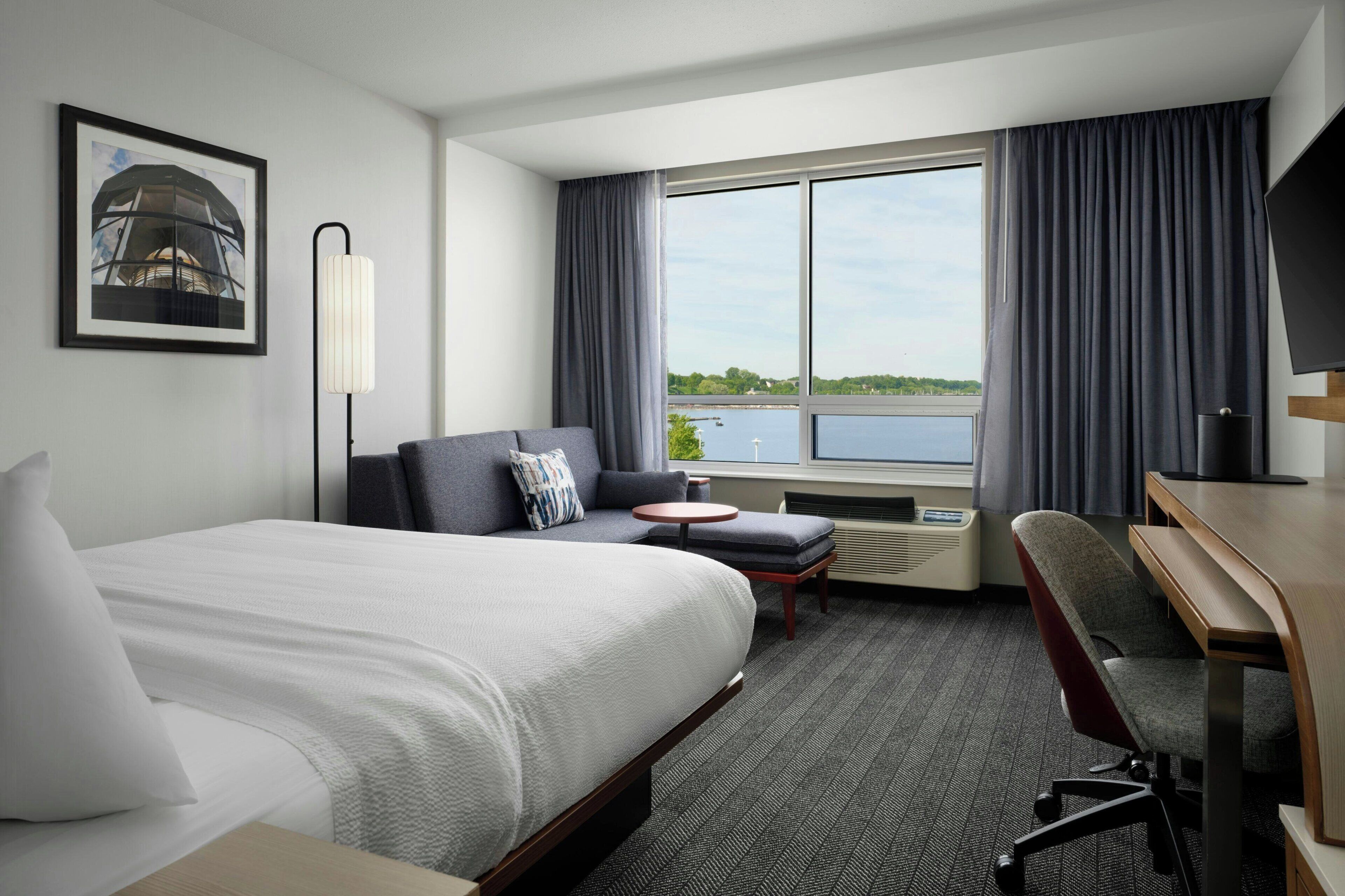 A hotel room with a bed, a sofa, a desk, a chair, a TV, a window with curtains, and a picture on the wall.