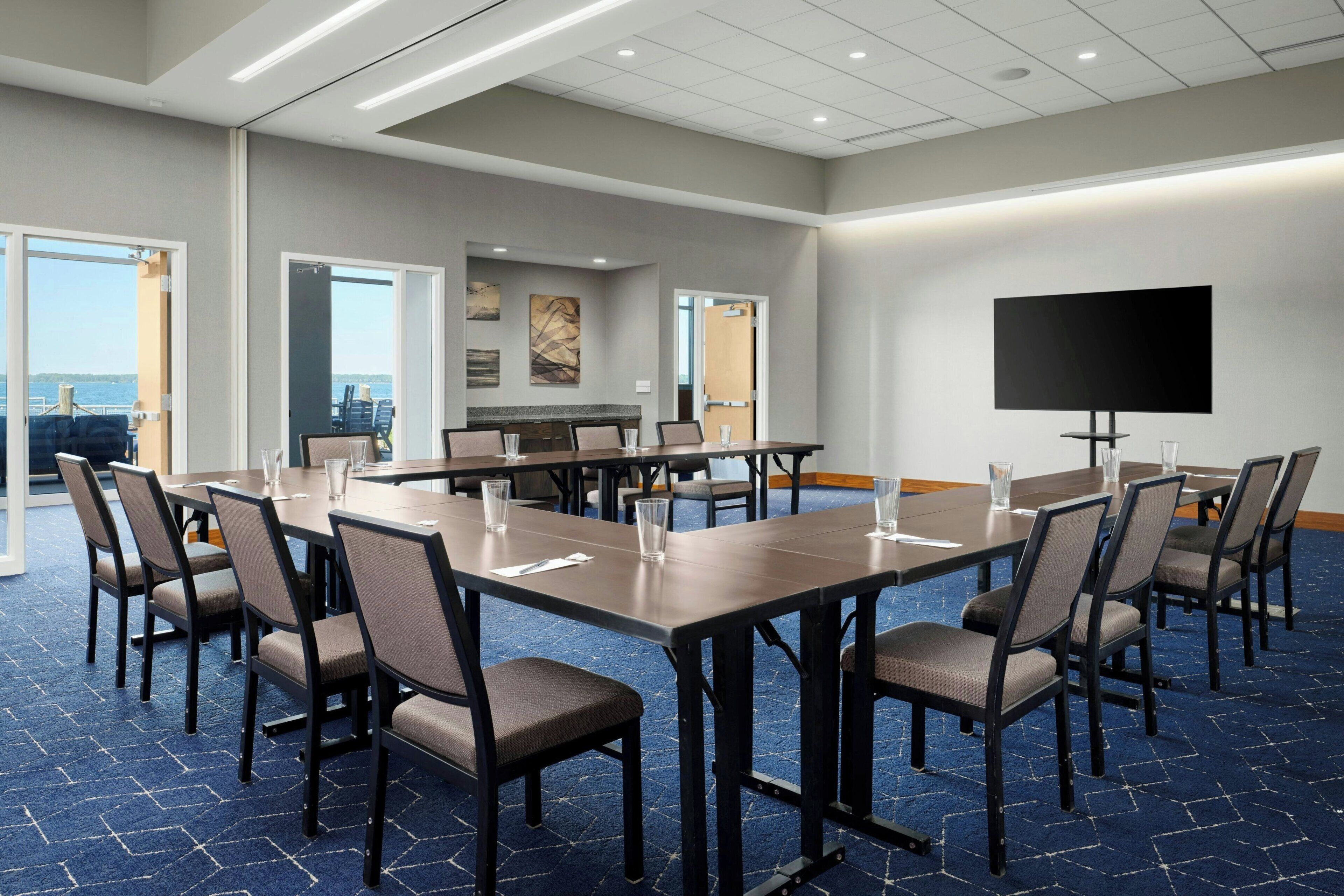 A conference room with multiple tables and chairs, a large flat-screen TV, and a view of the ocean through large windows.