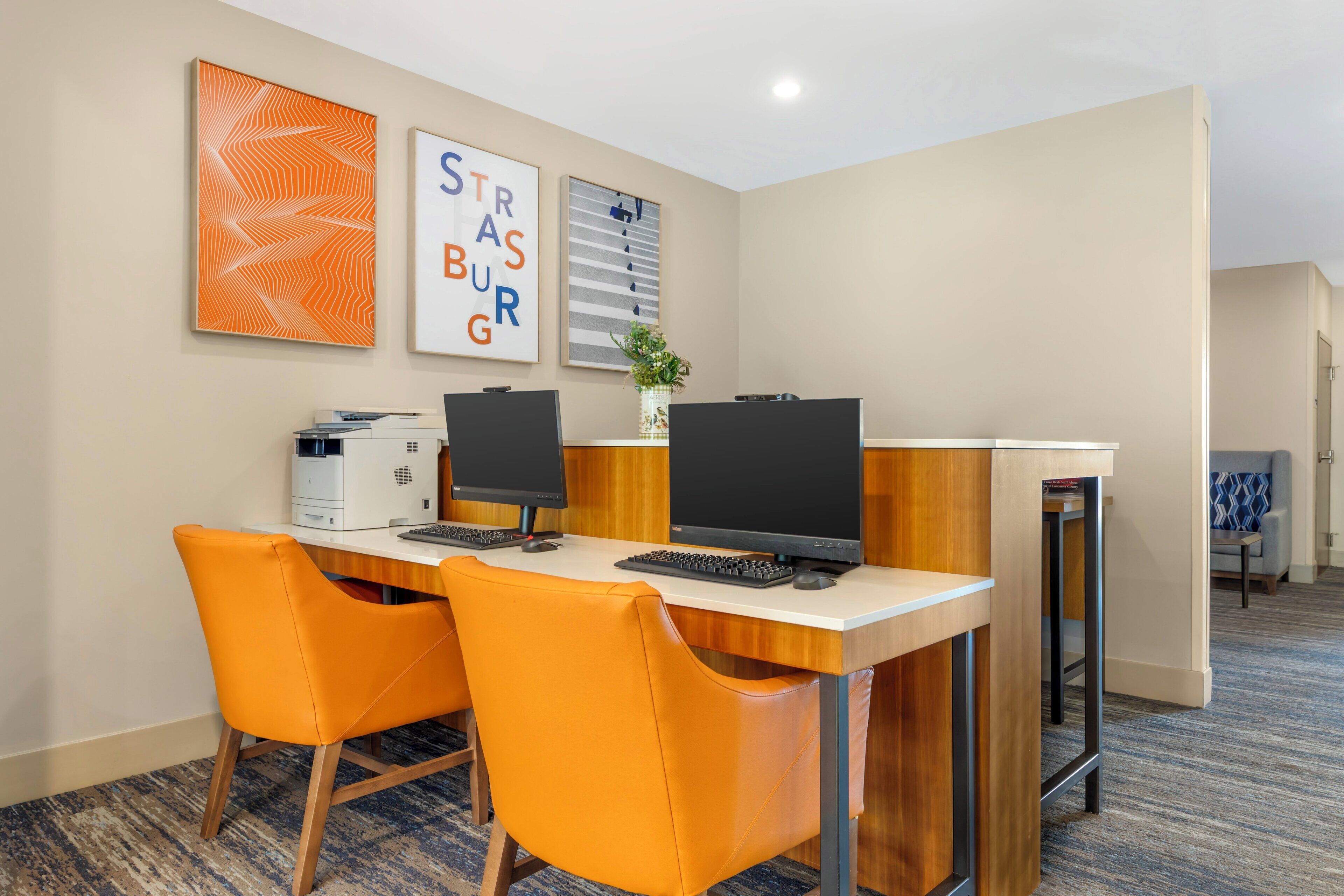 Habitación de hotel con un escritorio, dos monitores, una impresora y dos sillas naranjas.