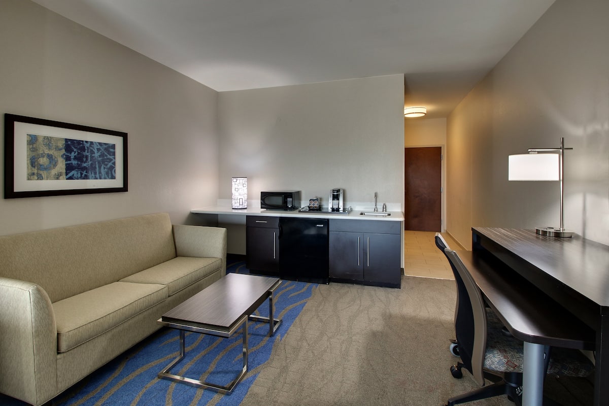 Habitación de hotel con sofá, una mesita, escritorio, microondas, cafetera y un cuadro en la pared.