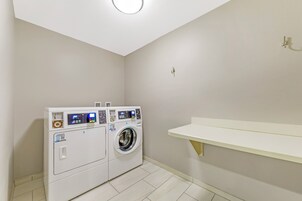 Un cuarto de lavado con dos lavadoras de carga frontal, un estante pequeño y una lámpara en el techo.