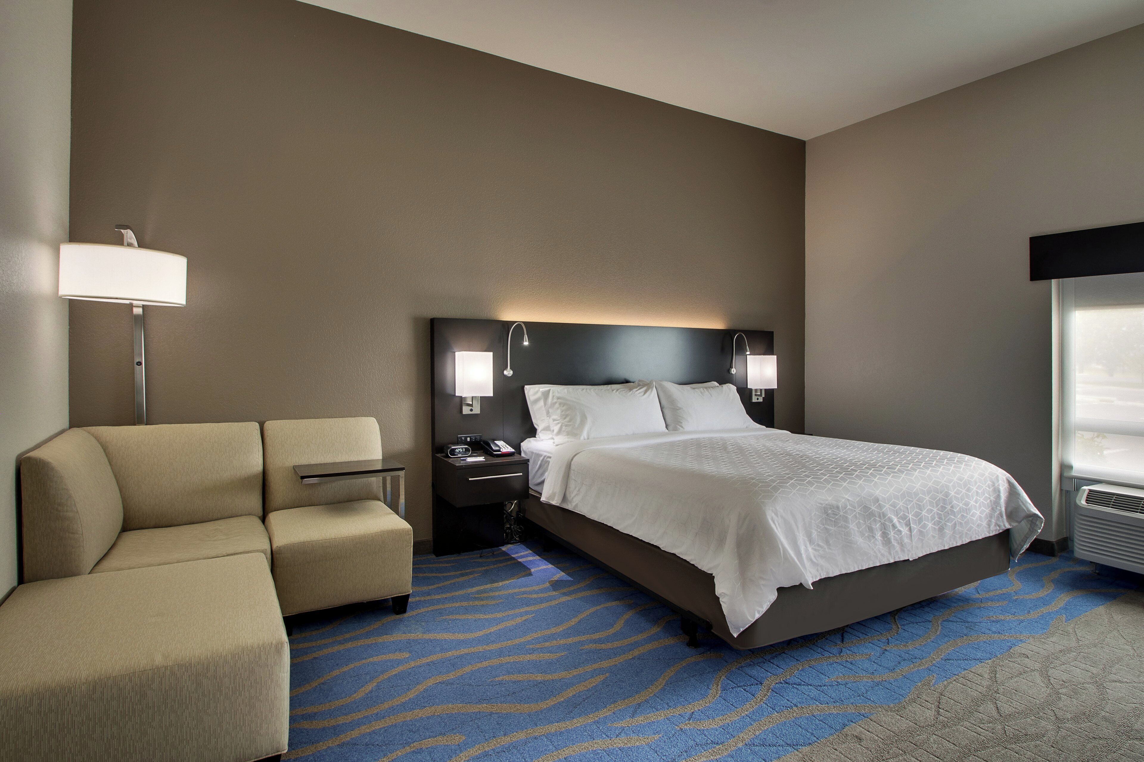 A hotel room with a bed, a sofa, a nightstand, a lamp, and a window.