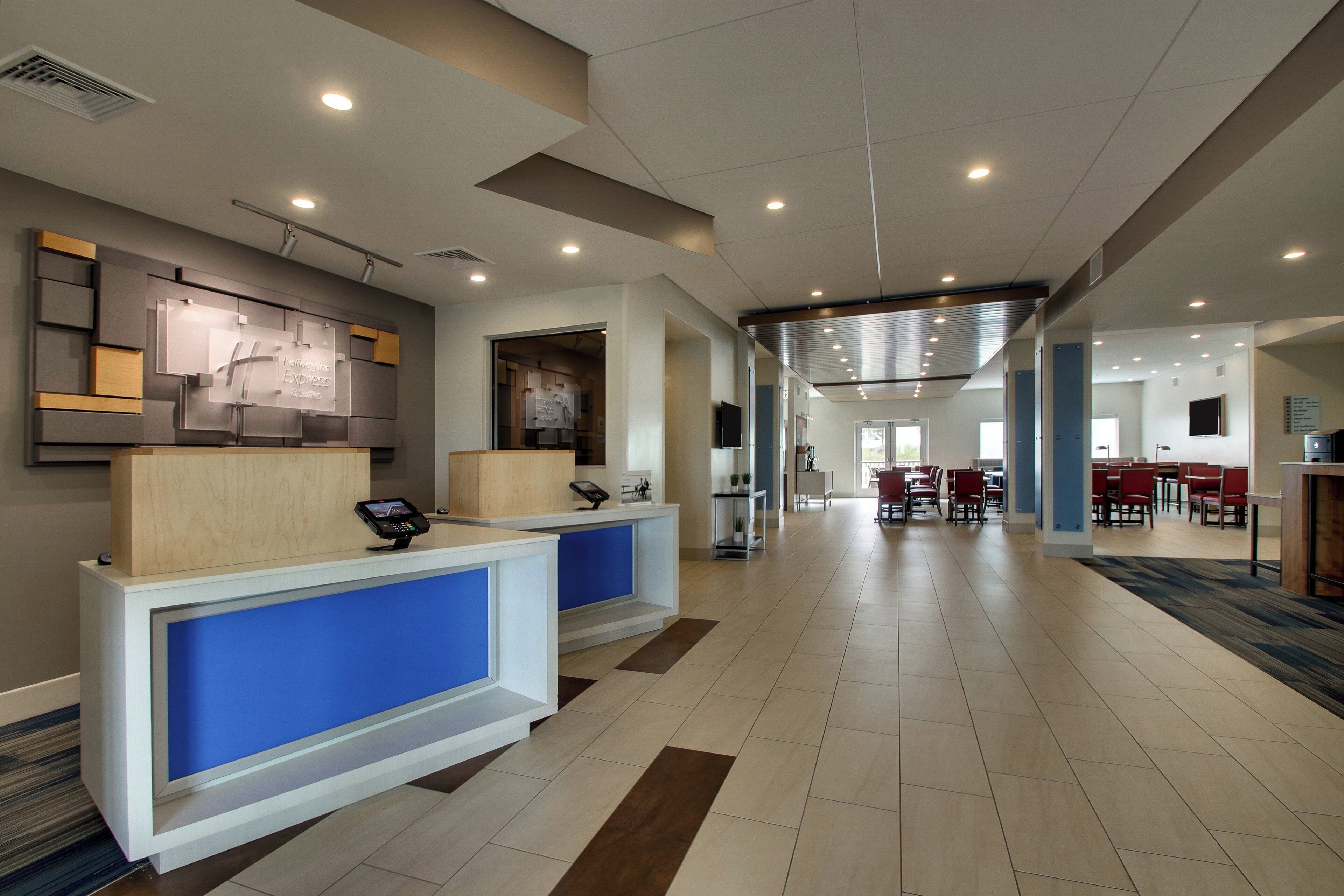 A hotel reception area with a modern design, including a reception desk, a digital information board, and a seating area with tables and chairs.