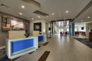 Een hotel receptie met een modern ontwerp, met een receptiebalie, een digitaal informatiebord en een zithoek met tafels en stoelen.
