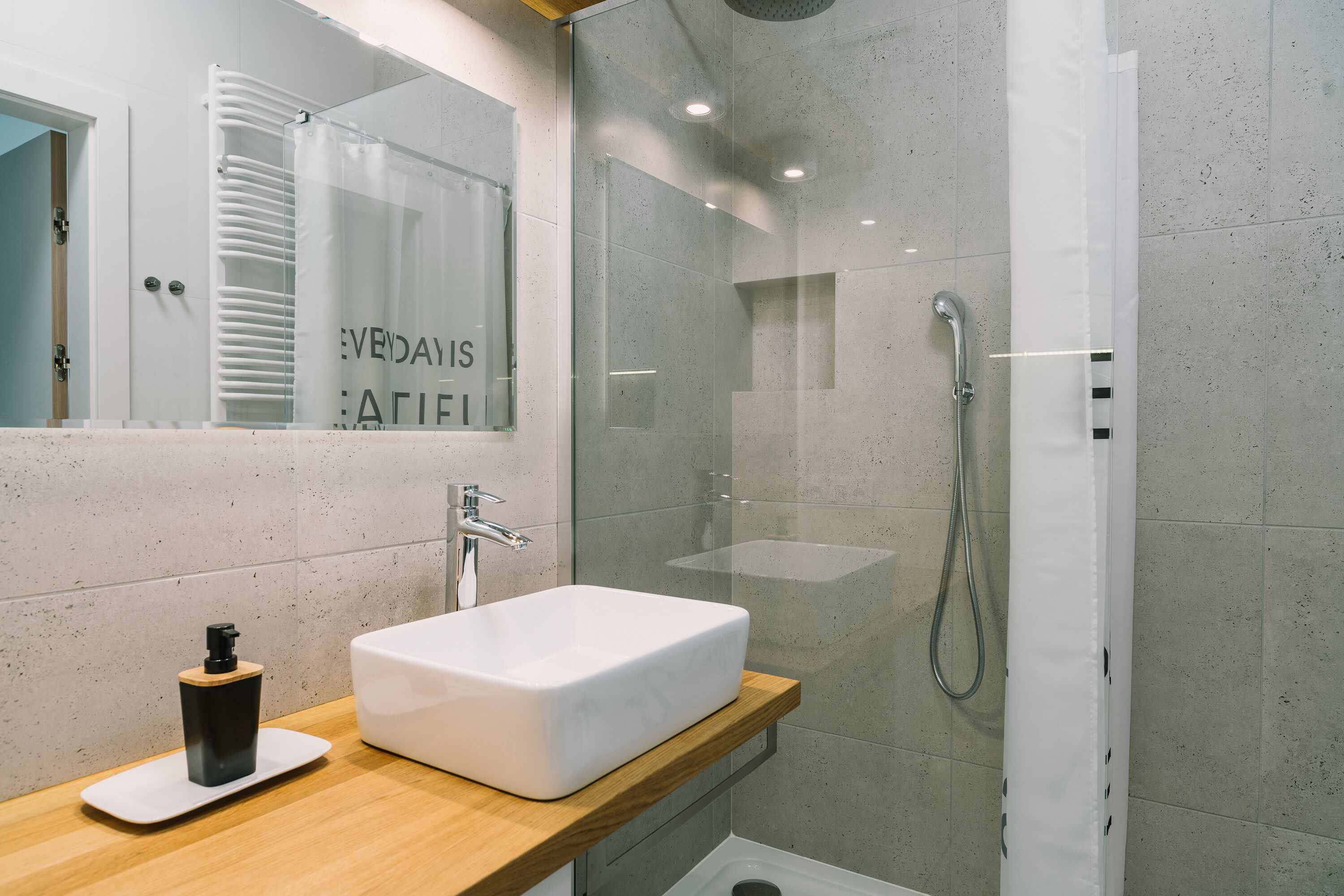 A modern bathroom with a large white sink, a wooden countertop, a glass shower enclosure, and a mirror with a frosted glass panel.
