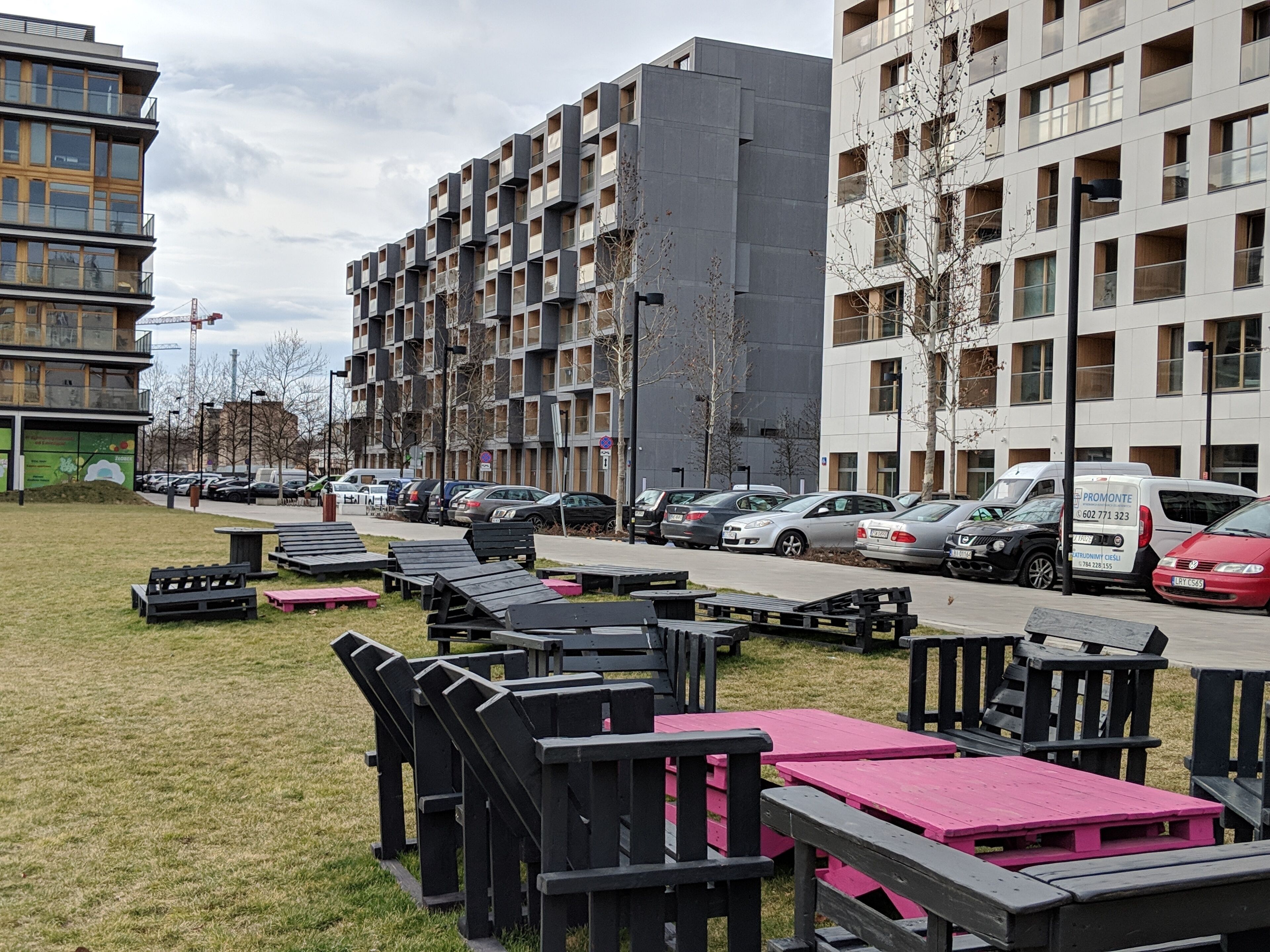 A modern apartment building with multiple floors and balconies, a parking area with several cars, and outdoor seating with tables and benches.