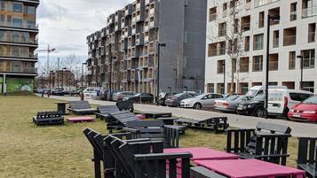 A modern apartment building with multiple floors and balconies, a parking area with several cars, and outdoor seating with tables and benches.