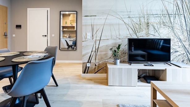 Een moderne woonkamer met een flatscreen televisie, een eethoek met blauwe stoelen, een houten kast en een wanddecoratie met een strandthema.
