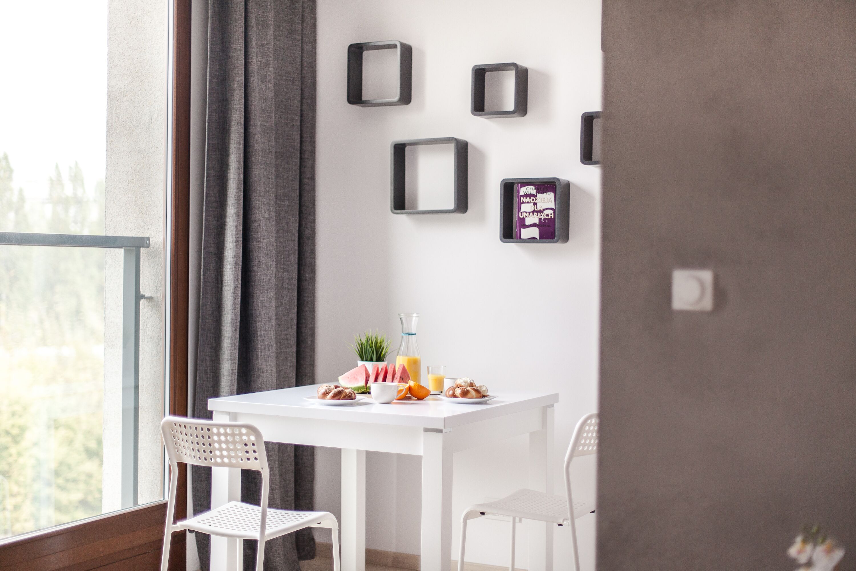 A modern room with a white table, two white chairs, a window with a view, and a wall with framed pictures and a small plant.