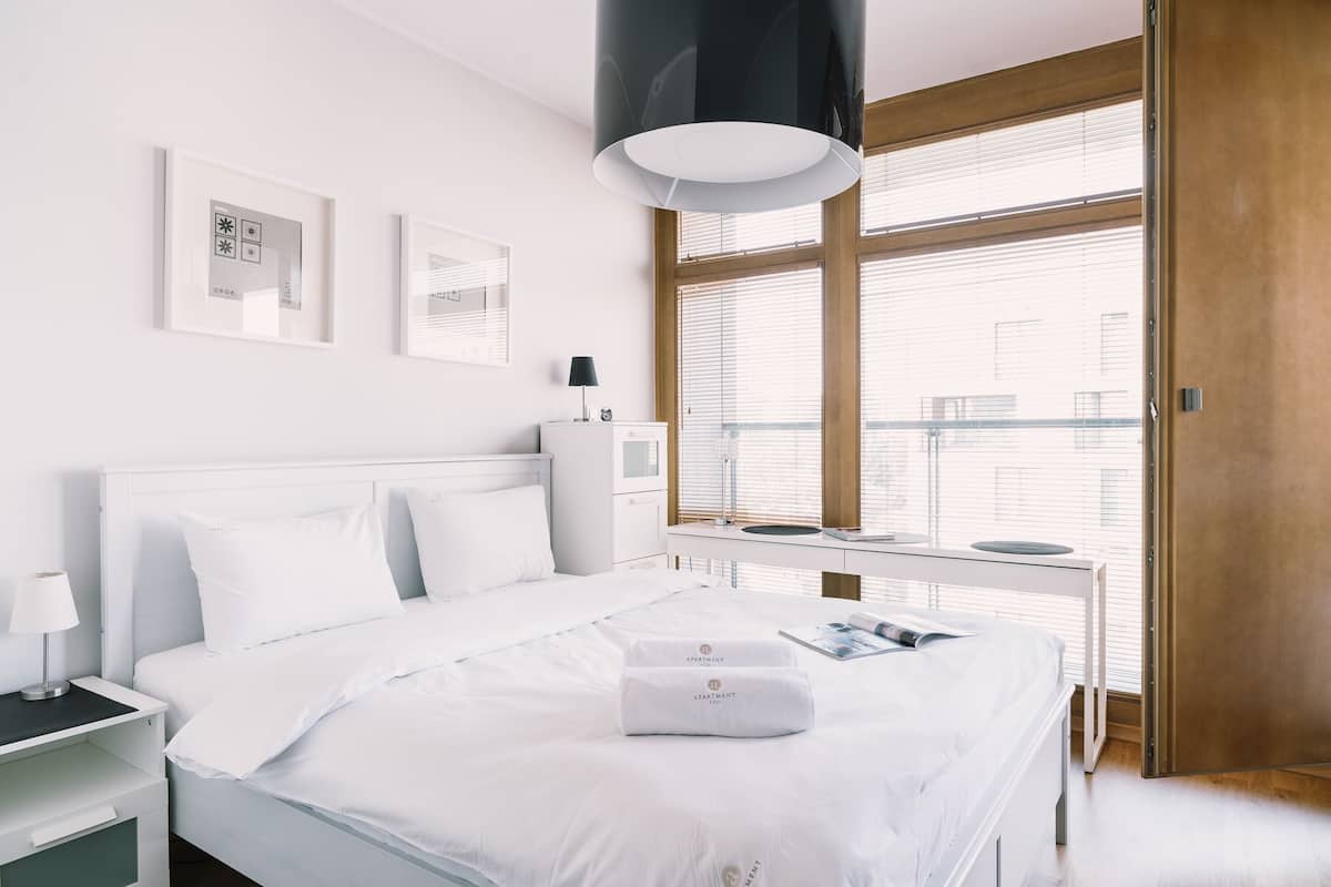 A modern hotel room with a neatly made bed, a bedside table with a lamp, a window with blinds, and a view of the city.