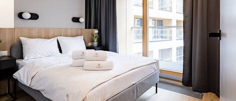 Quarto de hotel moderno com uma cama grande, uma mesa de cabeceira com luminária, uma planta em vaso e vista do edifício do lado de fora.