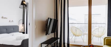 Ein modernes Hotelzimmer mit Bett, Fernseher, einem kleinen Tisch mit Kerze und einem Balkon mit Sitzgelegenheiten.