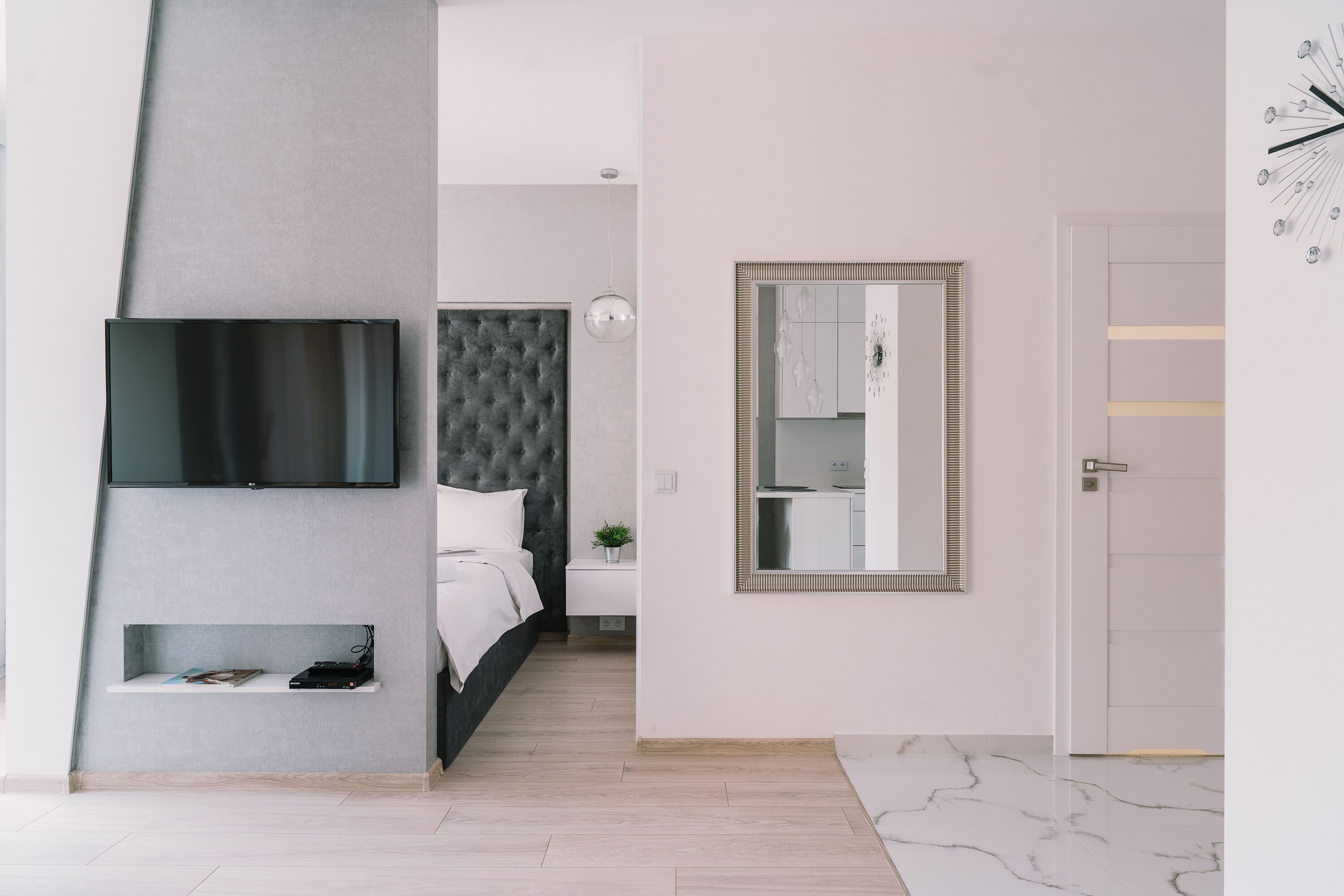 Una habitación de hotel moderna con televisor de pantalla plana, cabecero capitonado, mesita de noche con lámpara, espejo grande y suelo de mármol.