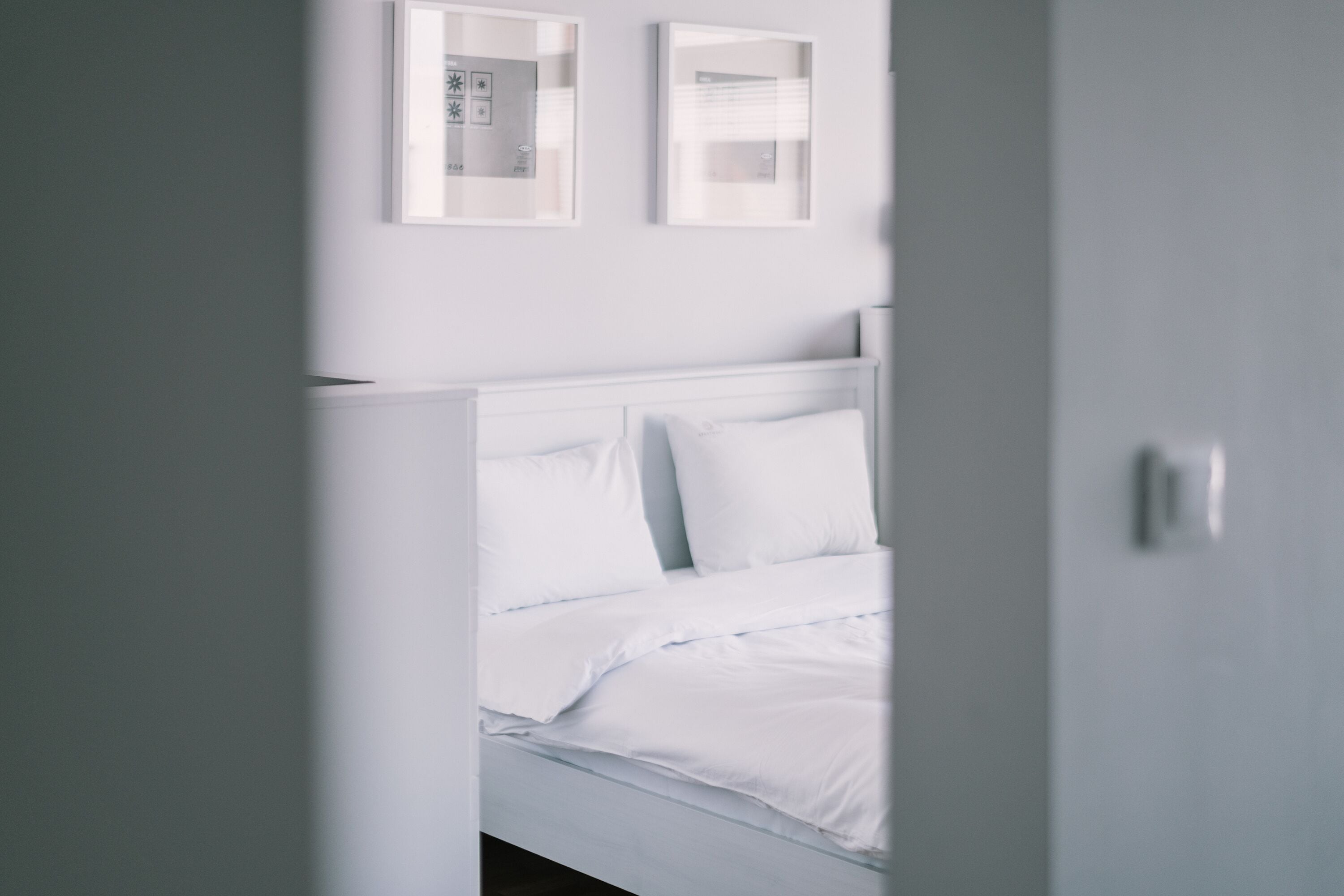 Een wit bed met hoofdeinde, een gedeeltelijk open deur en twee ingelijste foto's aan de muur.