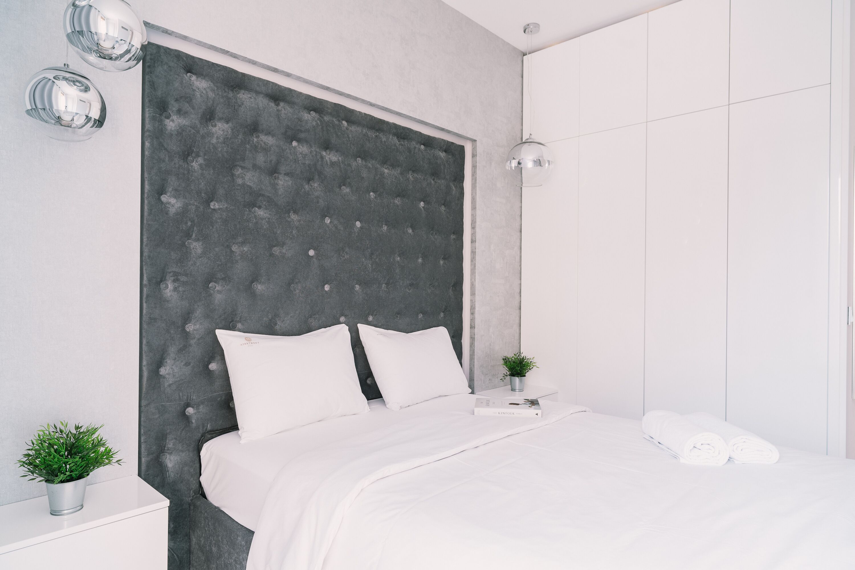 Een moderne slaapkamer met een gestoffeerd hoofdeinde, witte beddengoed, een nachtkastje met een plant en een hanglamp.