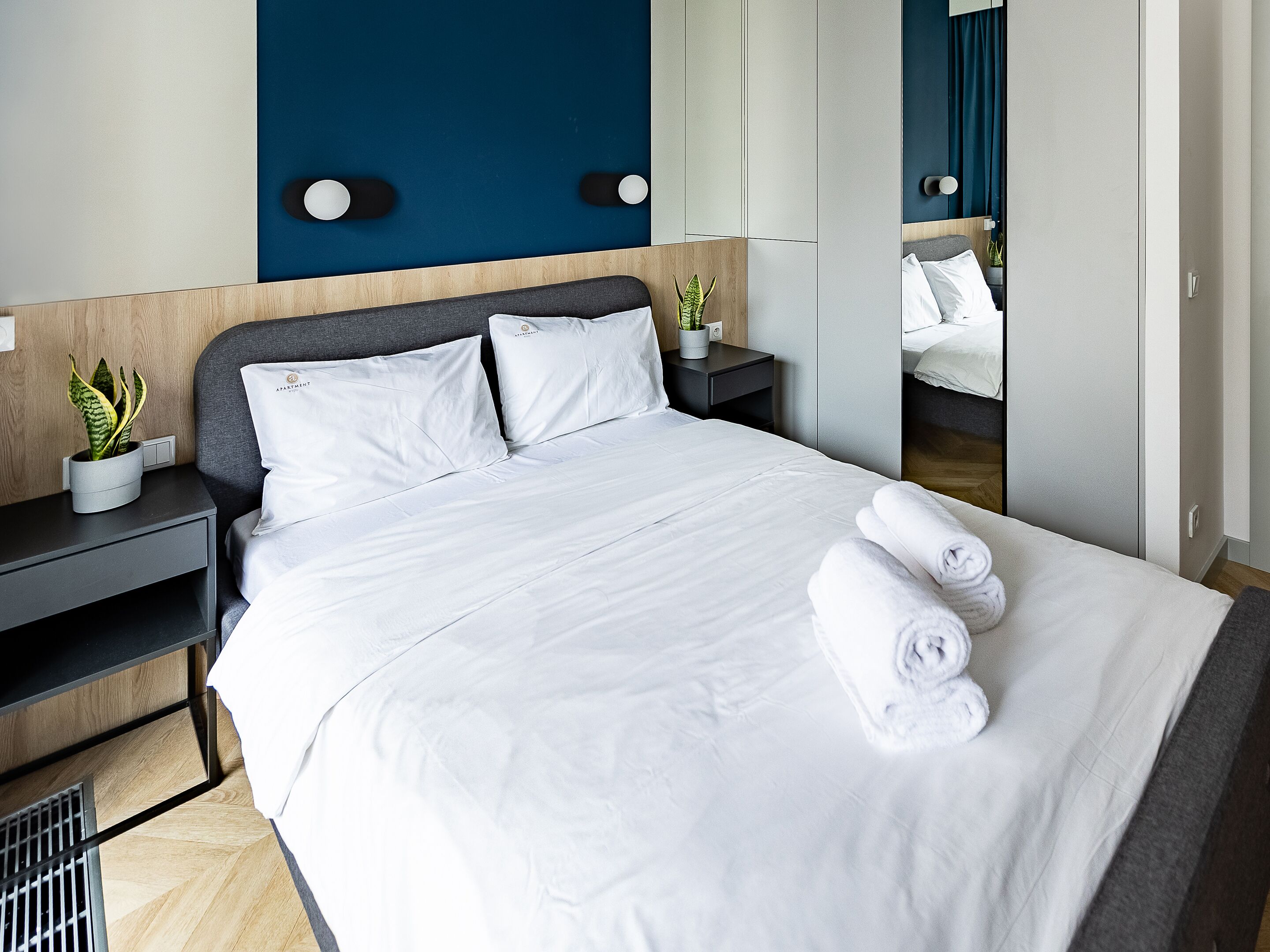 A neatly made bed with white linens and rolled towels, a bedside plant, and a modern headboard.