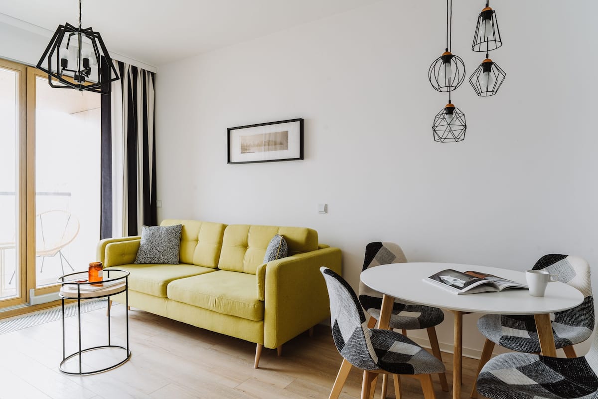 A modern living room with a yellow sofa, a round coffee table, and a dining area with a white table and patterned chairs.