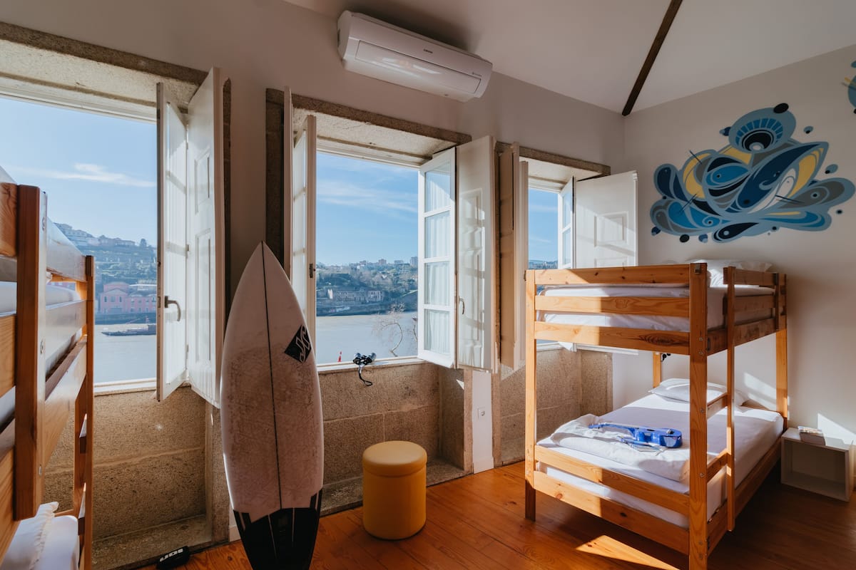 Ein Zimmer mit einem Etagenbett, einem runden Hocker und einem Fenster mit Blick auf eine Stadtlandschaft.