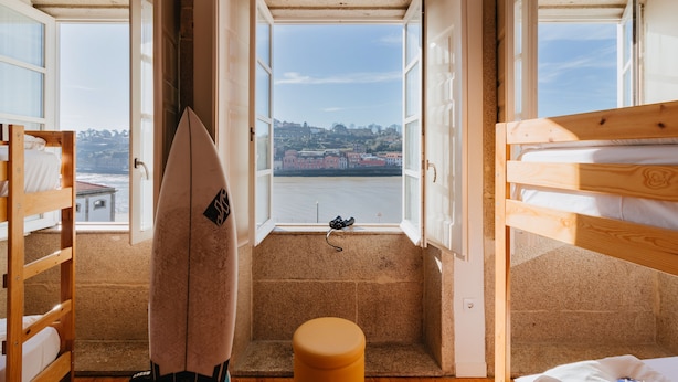 Um quarto com uma cama beliche, um banquinho pequeno e uma prancha de surf encostada na parede.