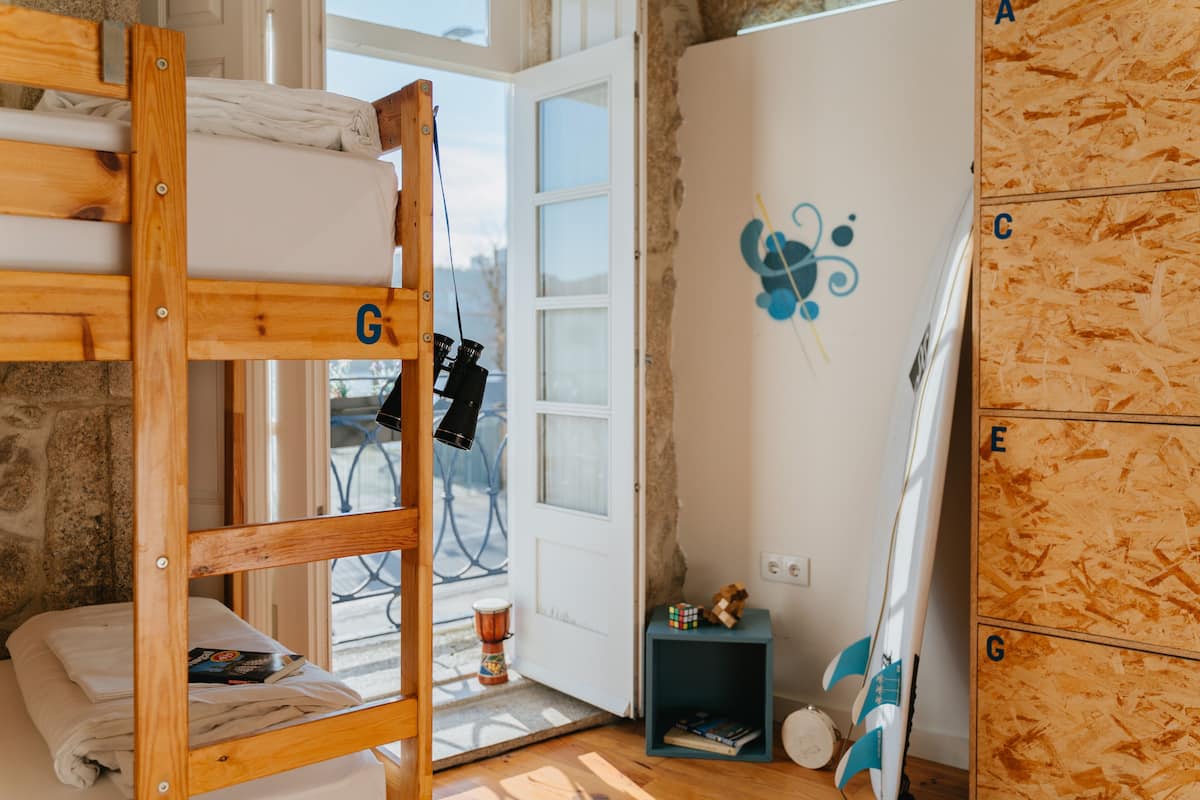 Ein Zimmer mit einem Etagenbett, einer kleinen Ablage mit Büchern, einem an der Wand angelehnten Surfbrett und einer zur Außenseite führenden Tür.