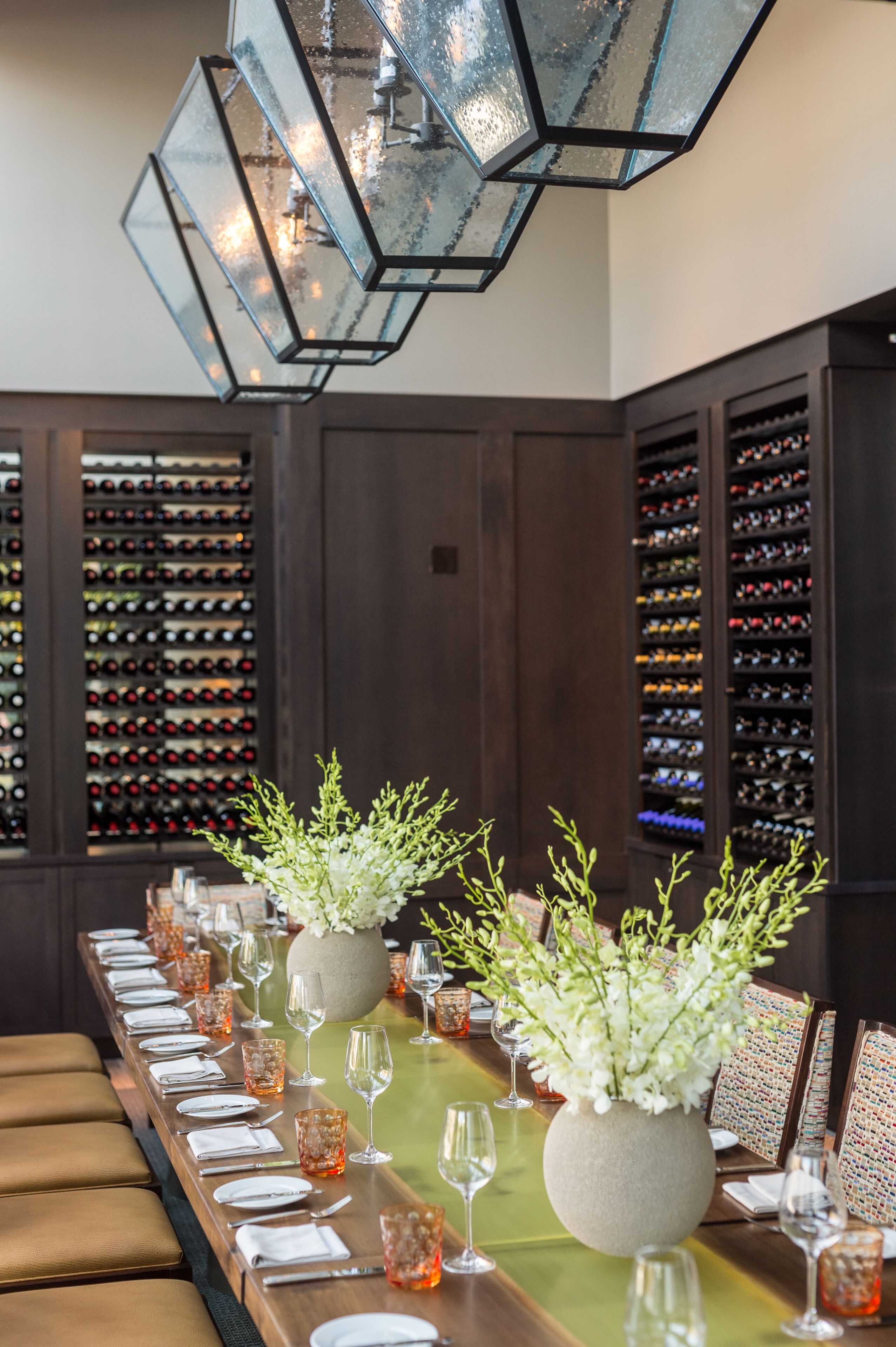 Una larga mesa de comedor dispuesta con platos blancos, cubiertos y cristalería, flanqueada por botelleros y una lámpara decorativa.