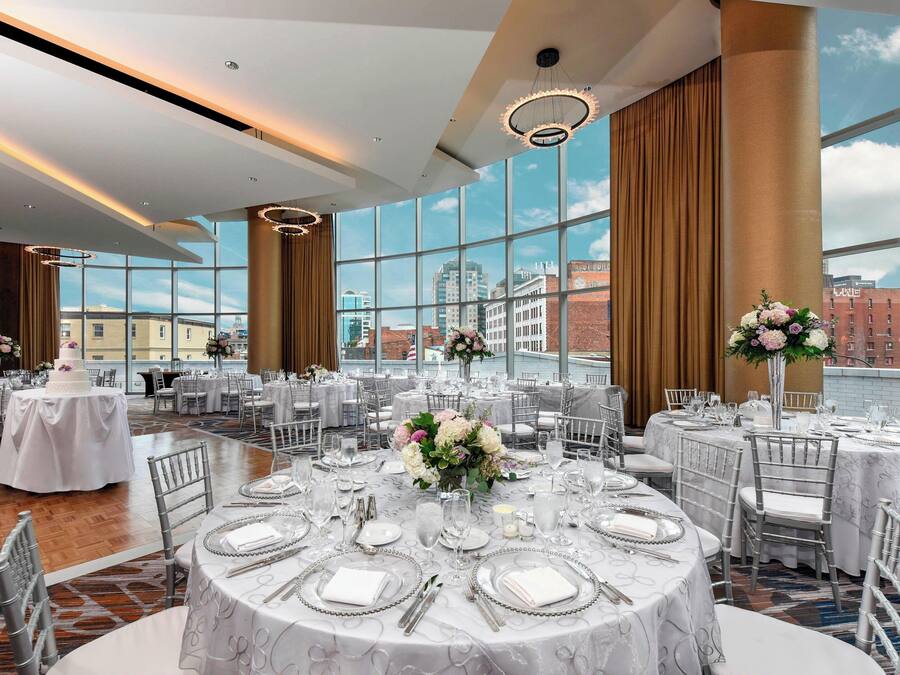 A spacious banquet hall with round tables set for a formal event, large windows offering a city view, and elegant lighting fixtures.