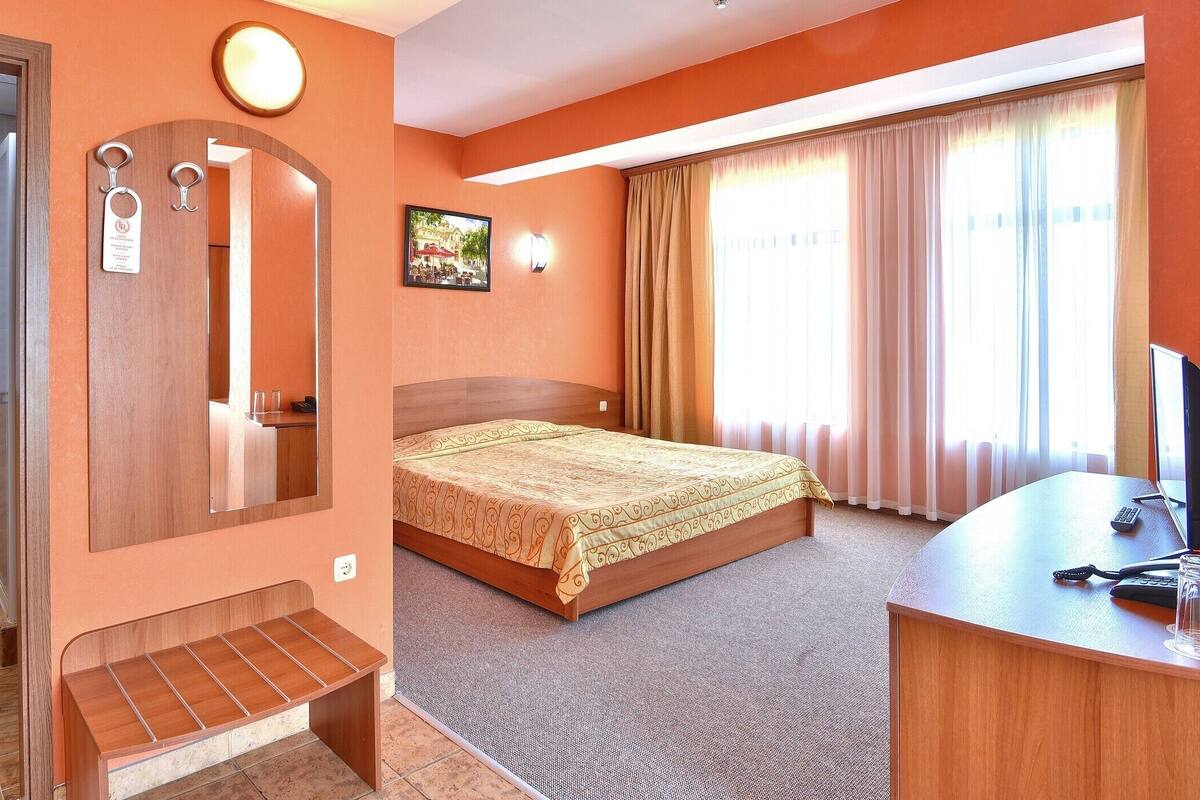 A hotel room with a bed, a desk with a computer, a mirror, and a bathroom with a shower.