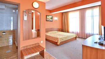 A hotel room with a bed, a desk with a computer, a mirror, and a bathroom with a shower.