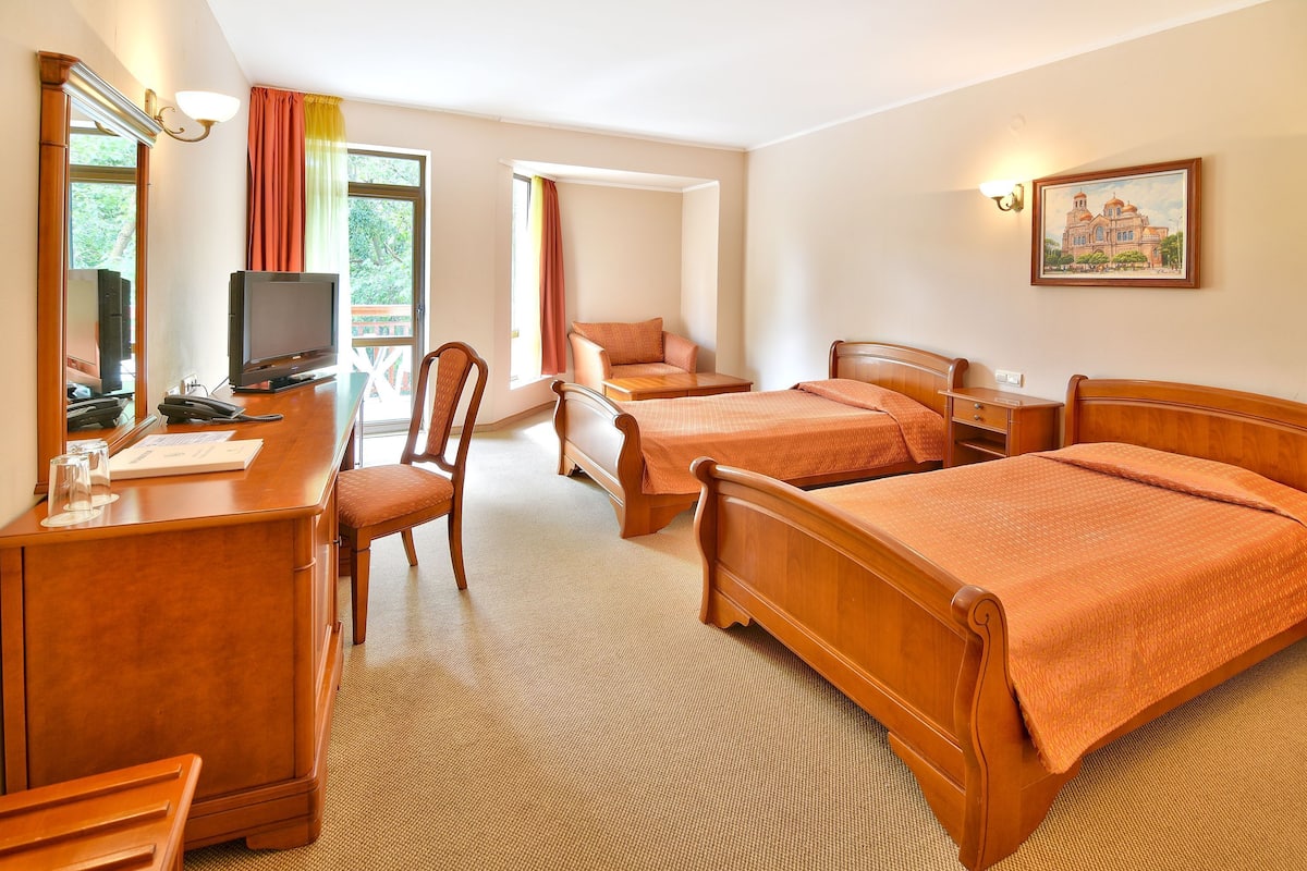A hotel room with a desk, television, two single beds, a chair, and a painting on the wall.