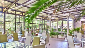Un espacio interior amplio con mesas y sillas de vidrio, rodeado de plantas en macetas y columnas.