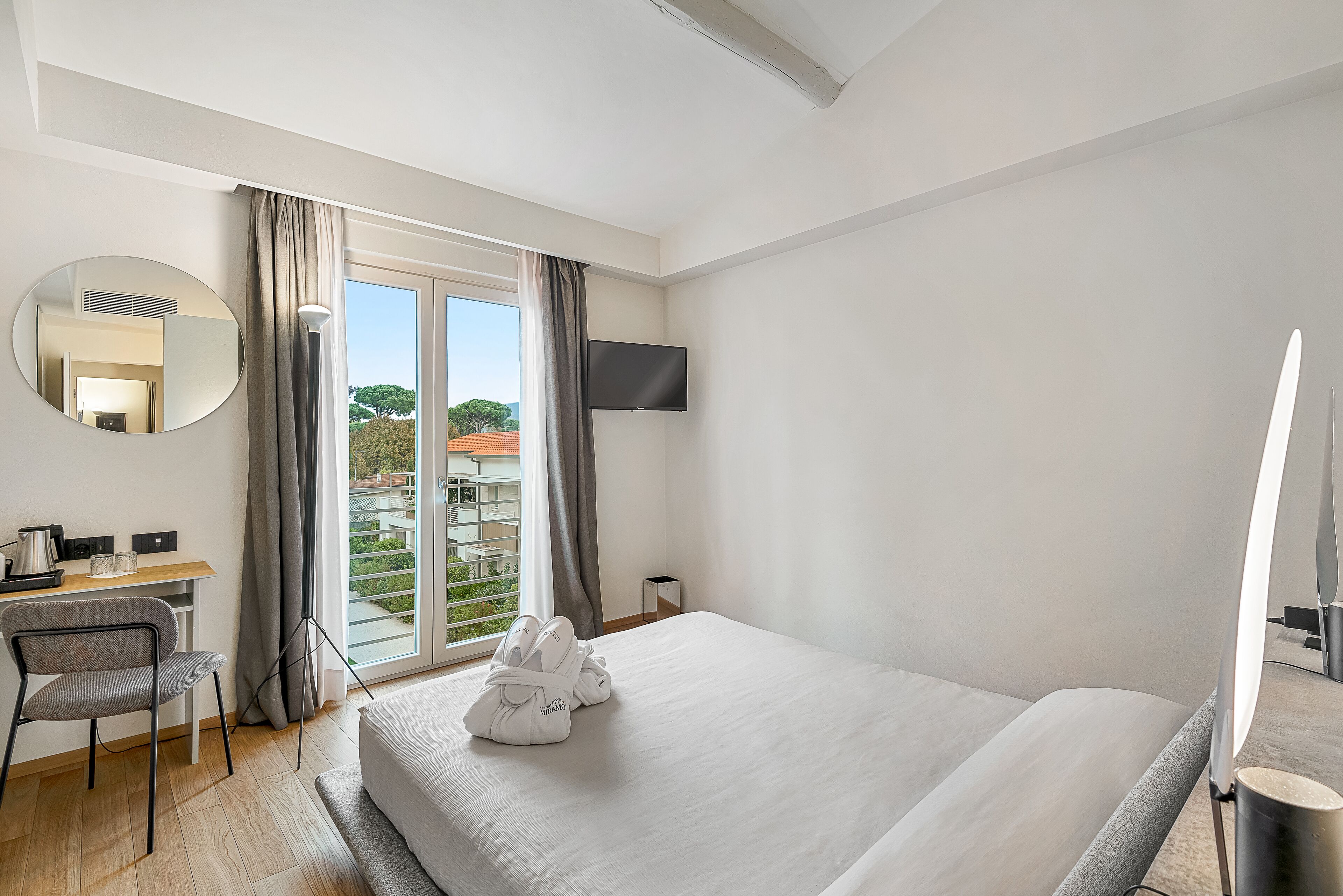 A hotel room with a bed, a desk with a chair, a TV mounted on the wall, and a window with curtains.