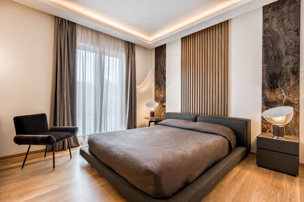 A modern bedroom with a large bed, a chair, a nightstand with a lamp, and a wall with vertical wooden slats.