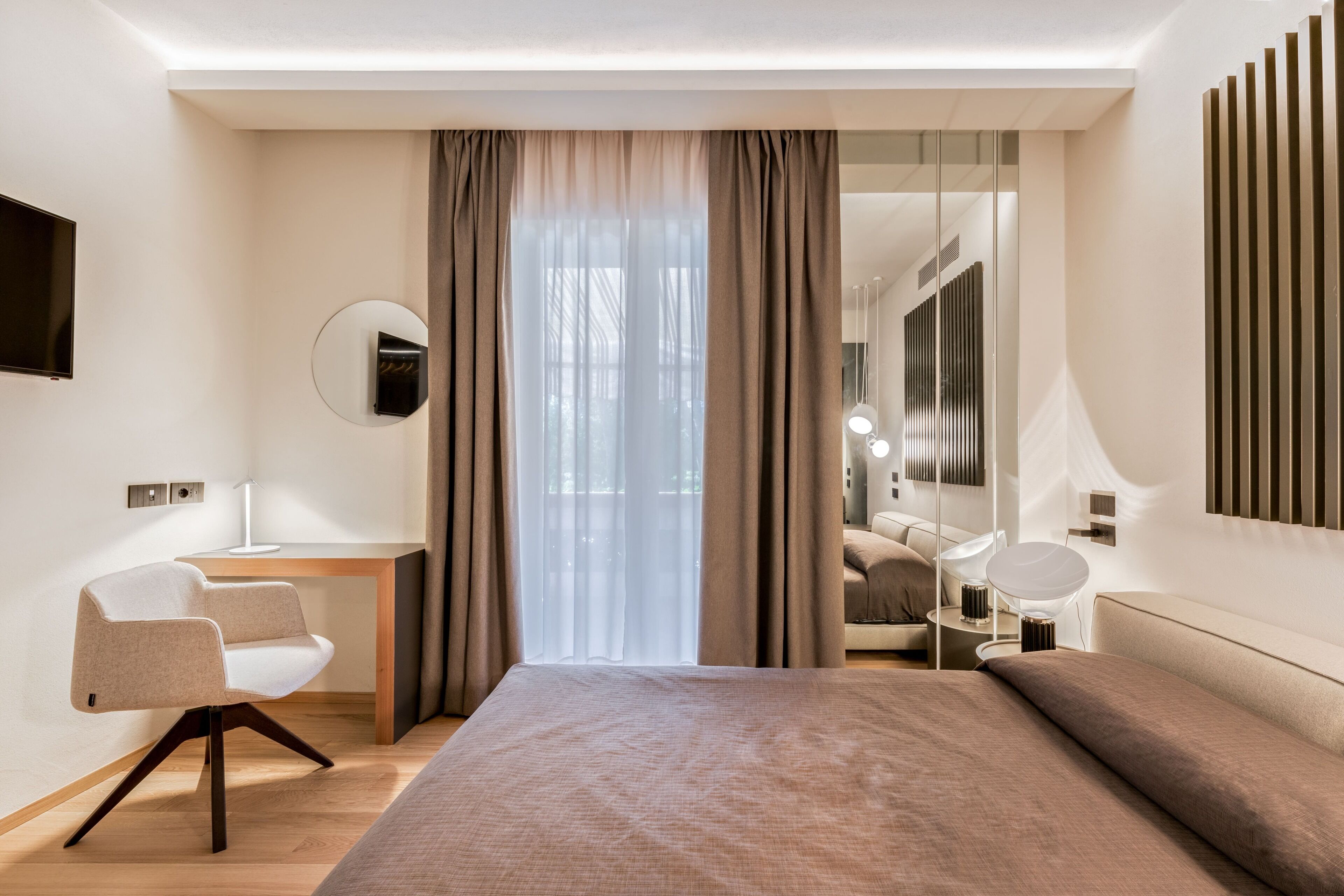 A modern hotel room with a large bed, a desk, a chair, and a mirror.