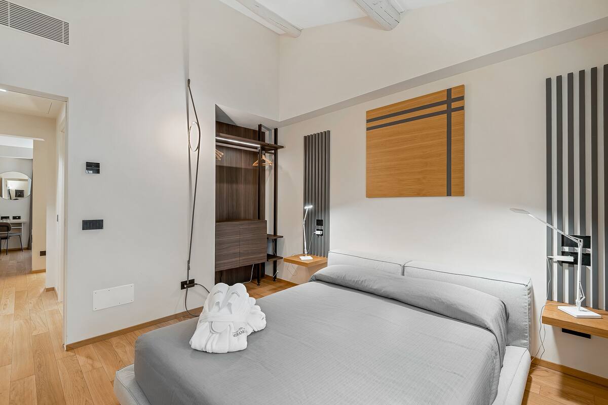A modern hotel room with a large bed, a wooden headboard, a bedside table with a lamp, and a wardrobe.