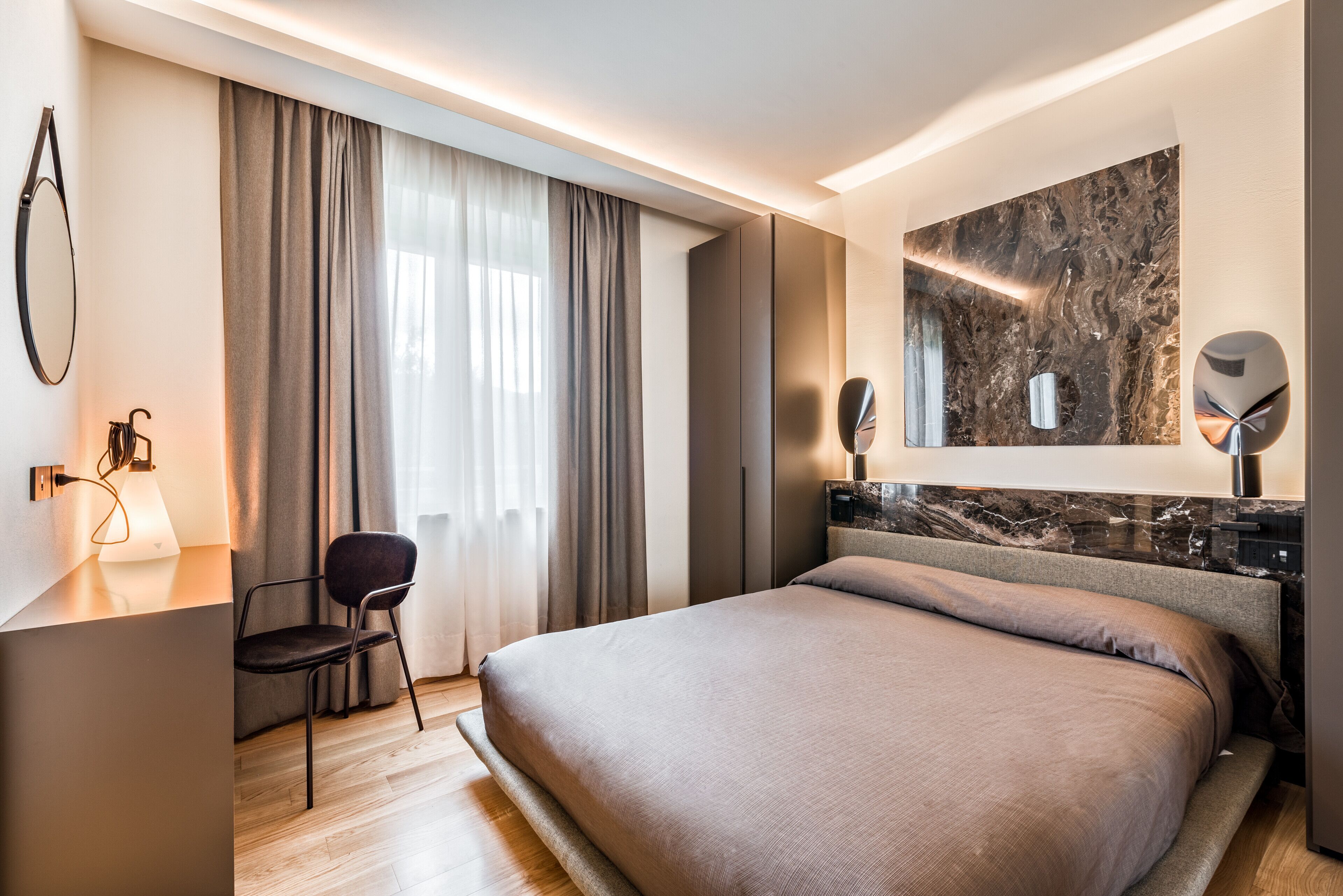 Ein modernes Hotelzimmer mit einem großen Bett, einem Schreibtisch mit Stuhl, einem großen Fenster mit Vorhängen und einer Wand mit Marmorverkleidung.