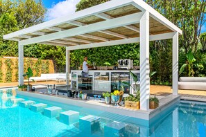 Zona de bar junto a la piscina con una pérgola, asientos y un barman.