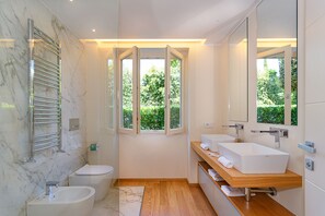 Un baño moderno con pared de mármol, un ventanal amplio, un lavabo blanco y un mueble de tocador de madera.