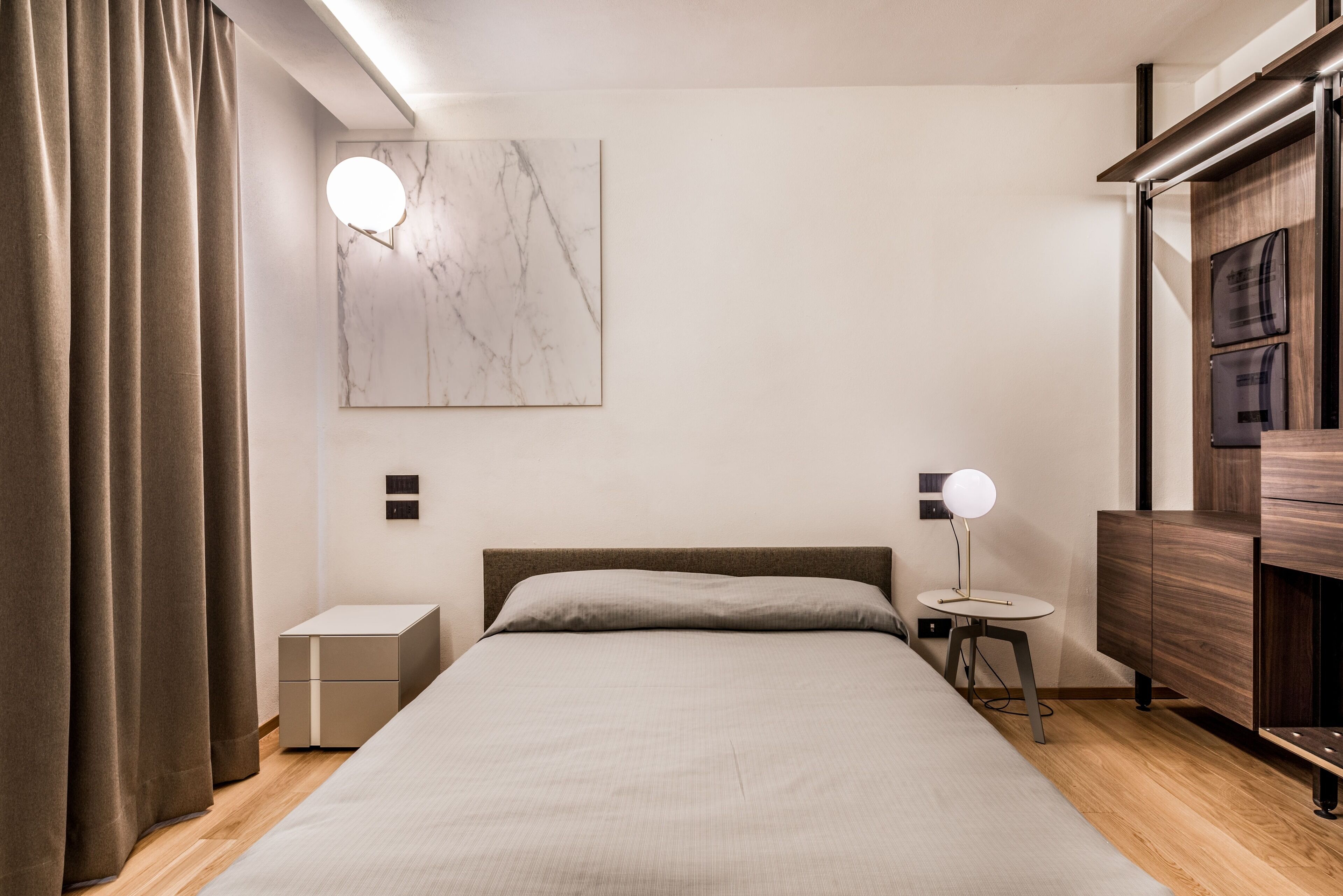Quarto moderno com cama, mesas de cabeceira, luminária na parede e cômoda de madeira.