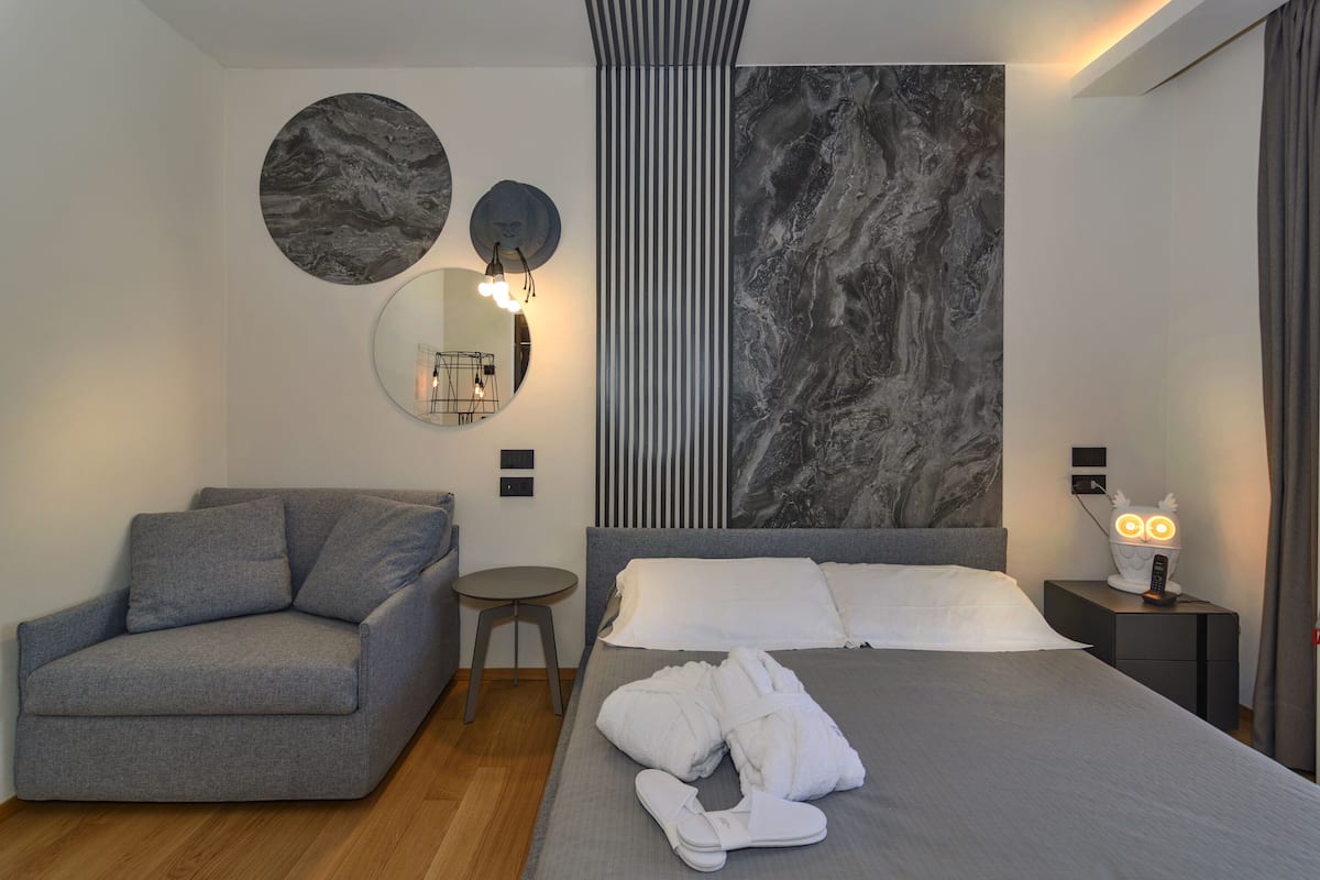A modern hotel room with a grey sofa, a round mirror, a bed with white bedding, and a wall with a marble-like pattern.