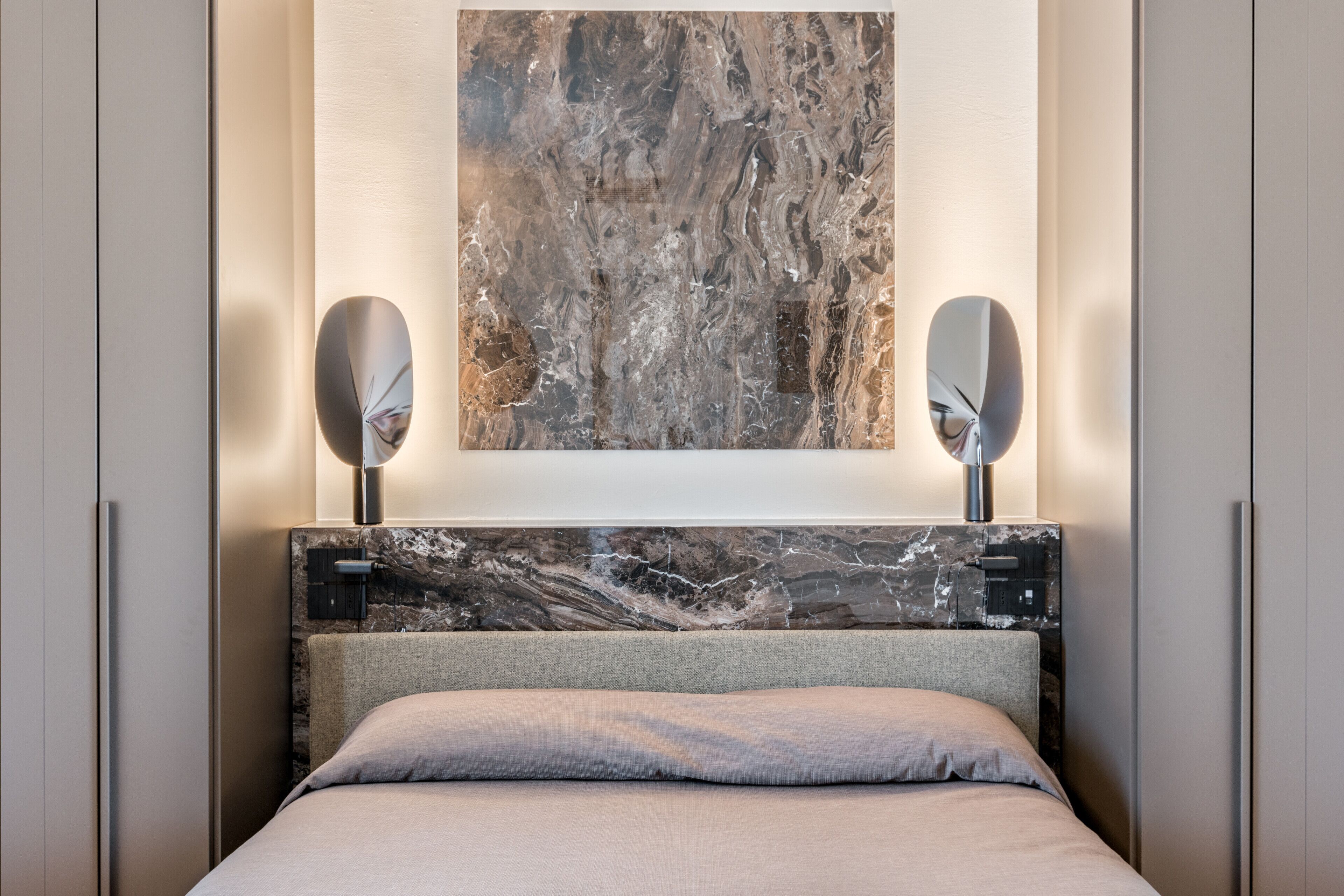 A modern bedroom with a marble headboard, a large abstract painting, and two wall-mounted mirrors.